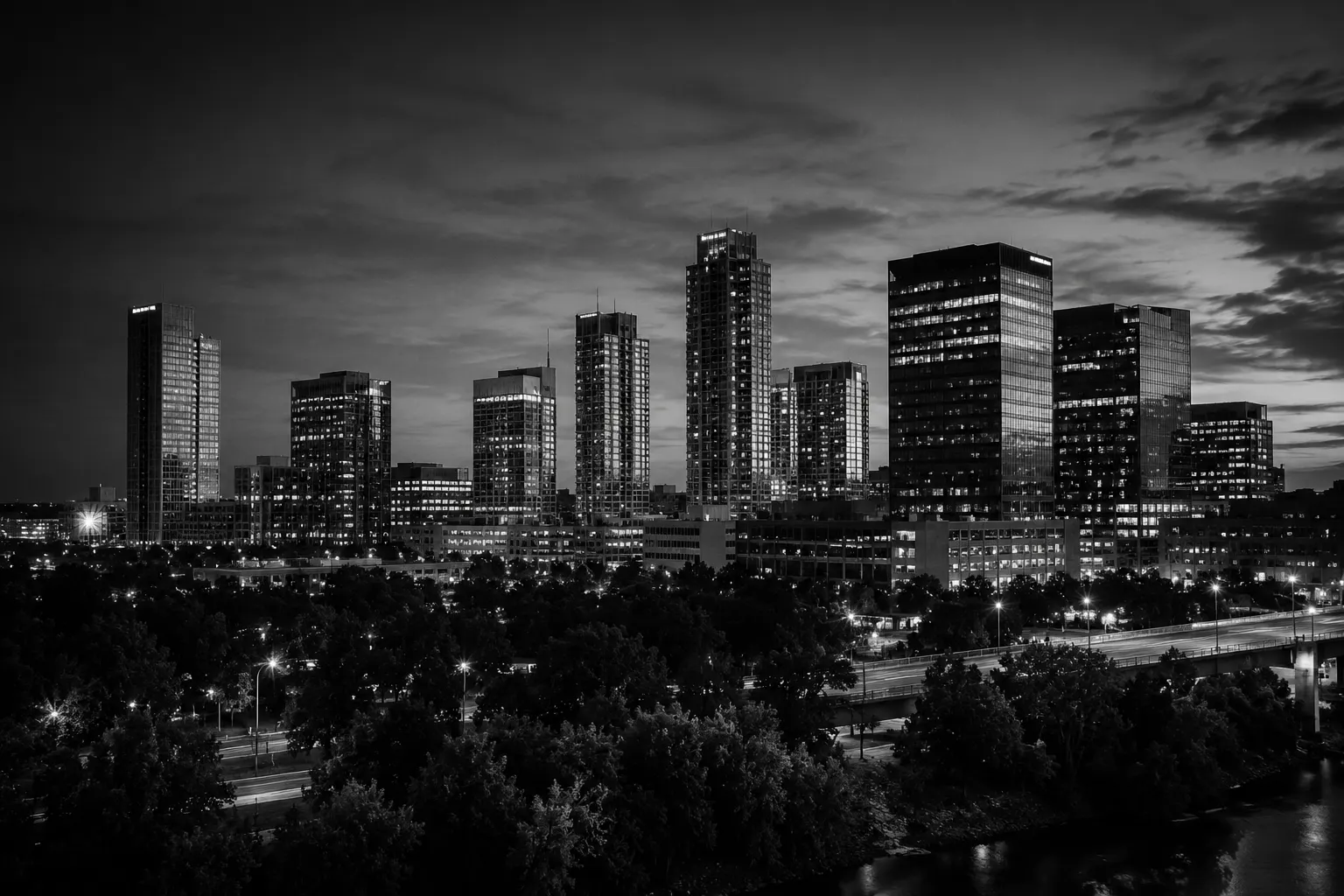 Northwest Arkansas skyline at dusk with illuminated high-rise towers rising above a tree-lined parkway, lit streets and a river crossing below.